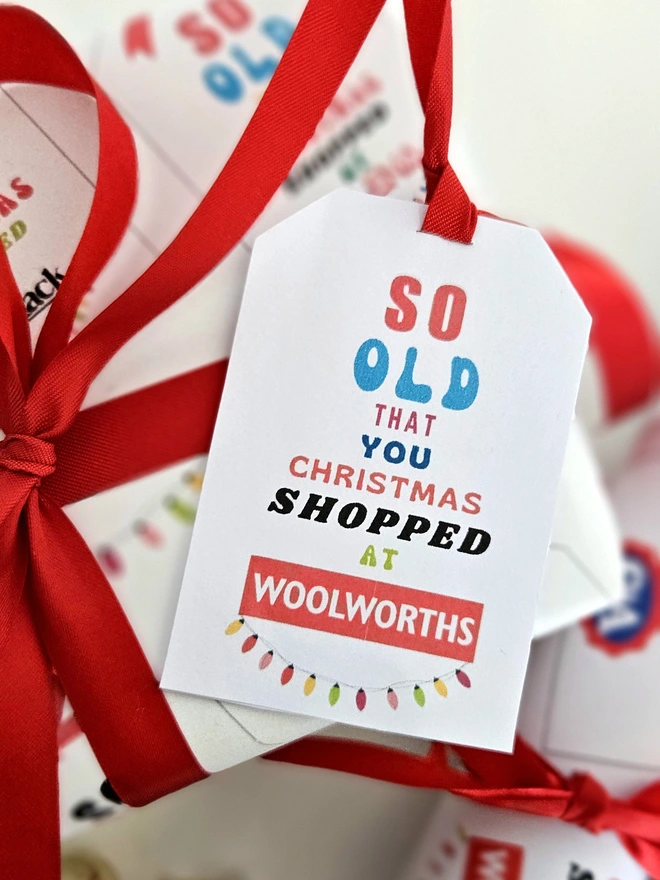 A tag for a present that says so old that you Christmas shopped at Woolworths written in brightly coloured text with an illustrated string of fairy lights. Attached to a present with a red ribbon.