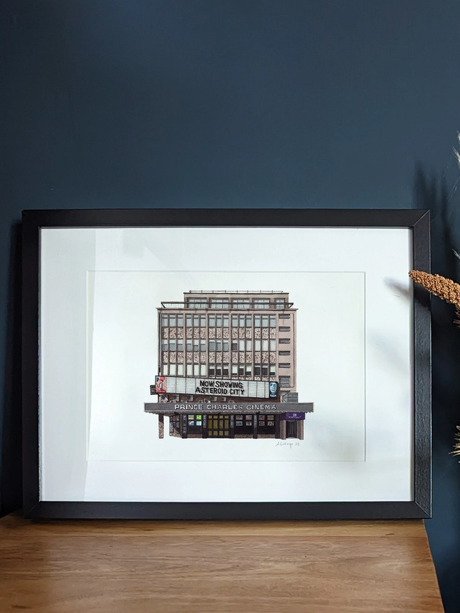 A framed watercolor print of the Prince Charles Cinema displayed on a wooden surface next to a vase of dried flowers.