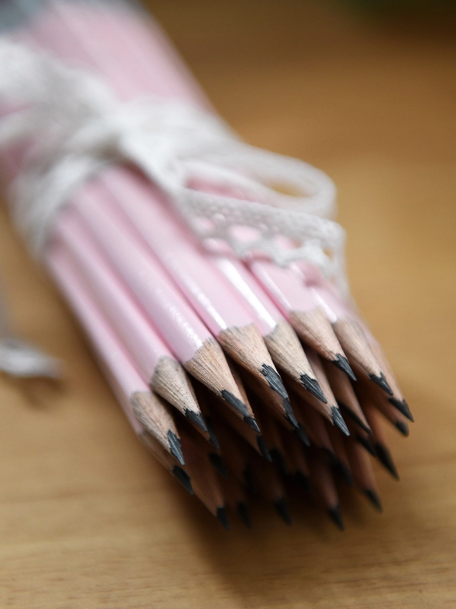 A bouquet of freshly sharpened pink pencils tied with a lace ribbon lays on a wooden desk.