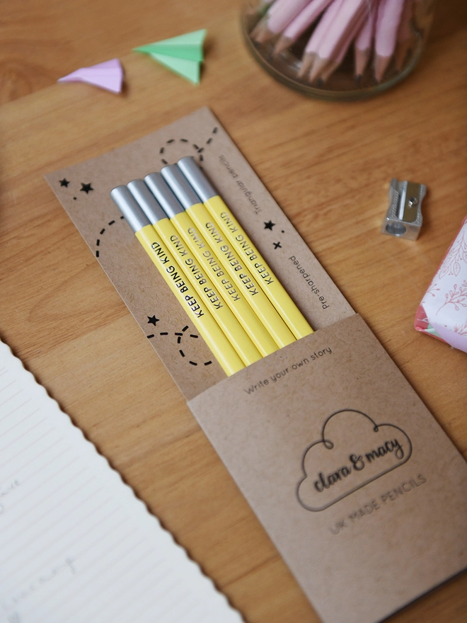 Five yellow pencils with the words Keep Being Kind along the side of each one, are tucked into cardboard packaging on a wooden desk.