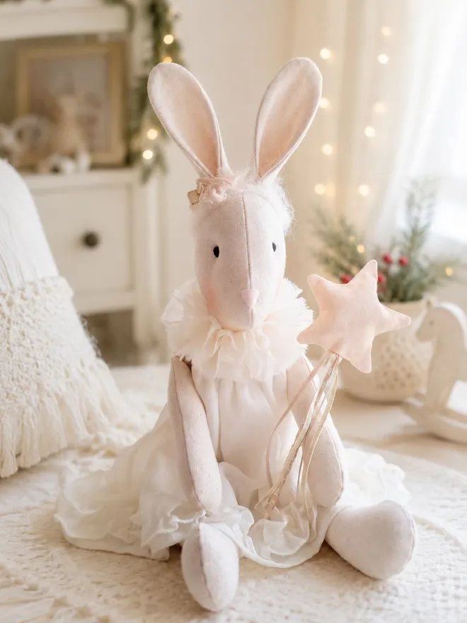 Heirloom linen bunny Veronica Christmas angel. She wears a chiffon floaty dress and traditional choir collar. She has a alpaca yarn headband with lace and ribbon rosettes and is holding a gold satin star wand on a wooden stick.