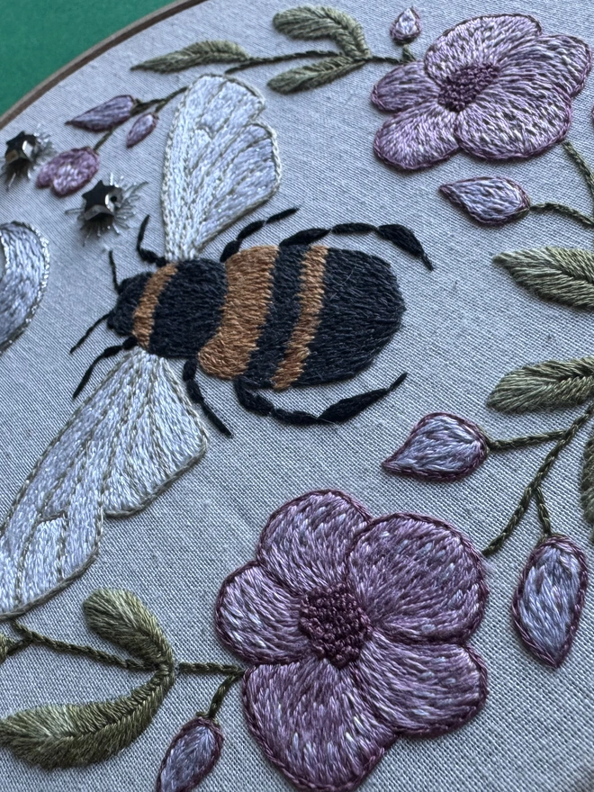Embroidery hoop containing a stitched bee pink flowers green leaves