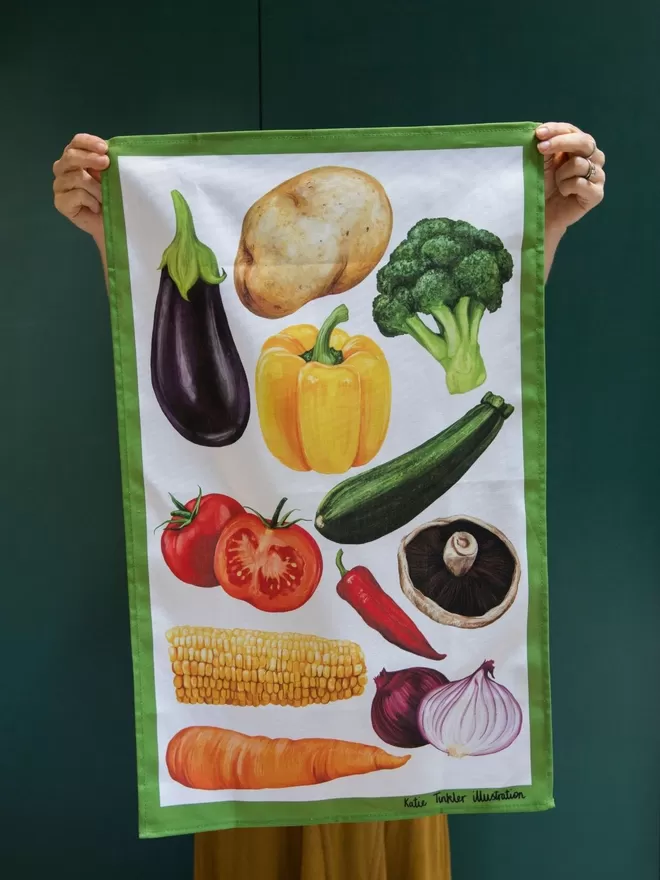 Vegetable Themed Tea Towel seen held up in front of a green wall.