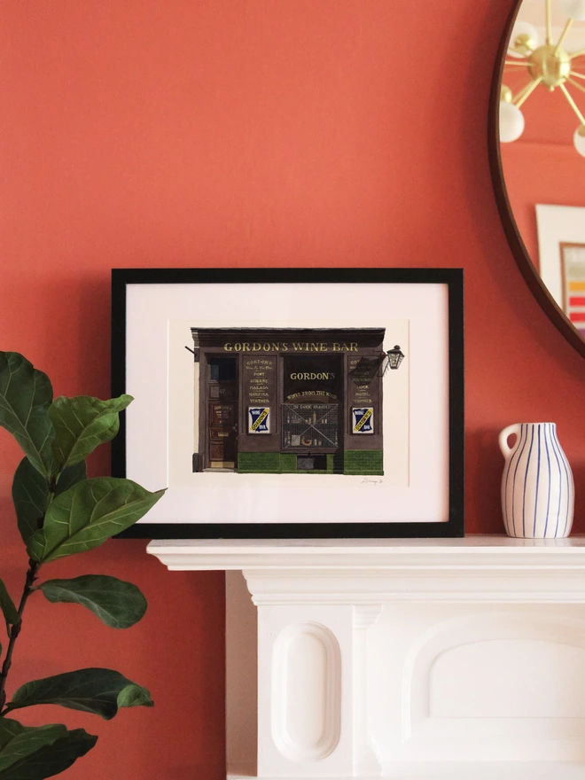 A framed watercolor print of Gordon's Wine Bar sitting on a white mantelpiece against a pink wall.