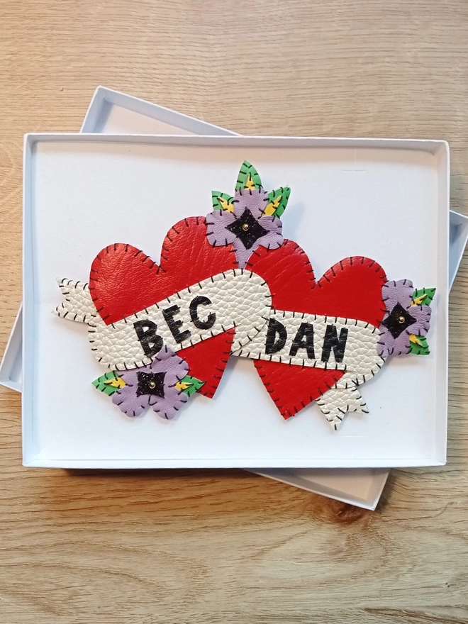 A large brooch of two red leatherette hearts intertwined with purple flowers, and white scrolls with the names Bec and Dan hand stitched in black lettering