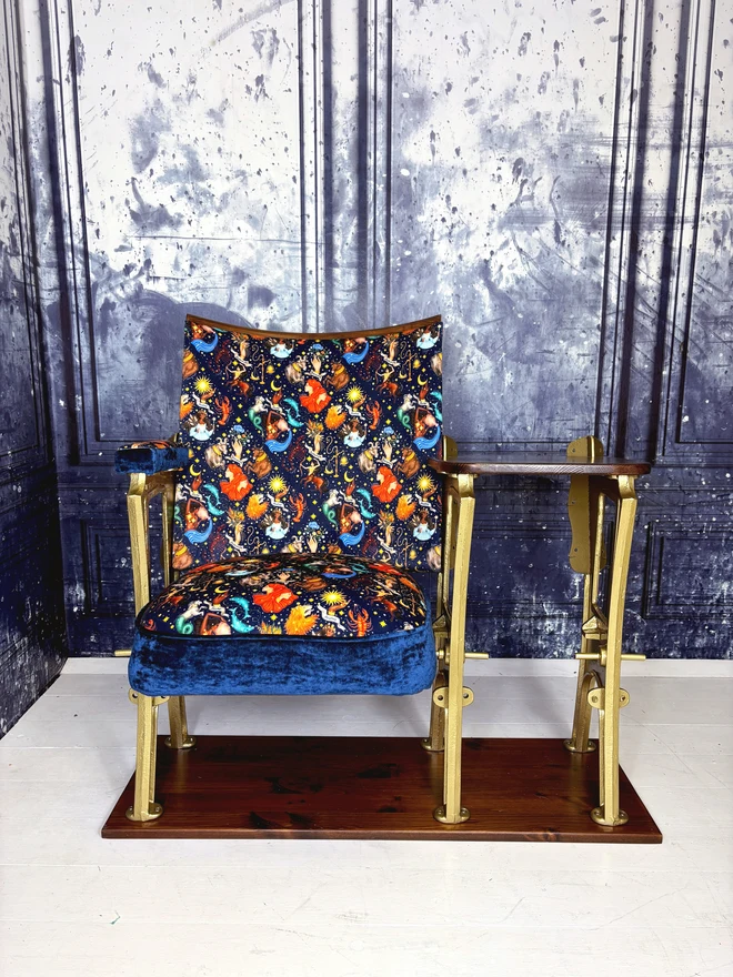 a vintage cinema seat with side table.  The flip down part of the seat is open.  The chair is upholstered in a navy velvet with astrology illustrations on it.  The chair is in front of a blue marbled wall. 