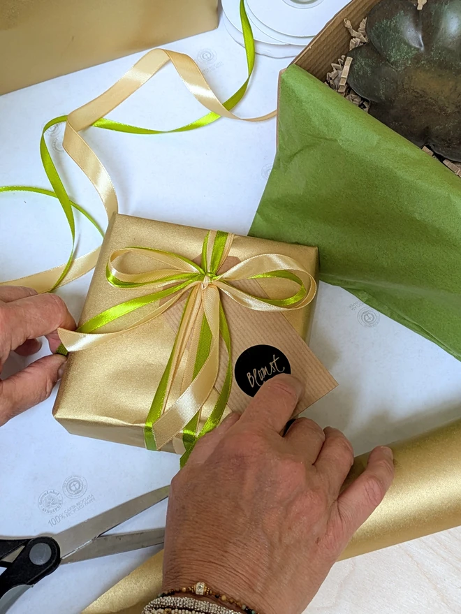 Giftwrapped order from Blomst, gold recycled paper with hands tying a bow in gold and green satin ribbon