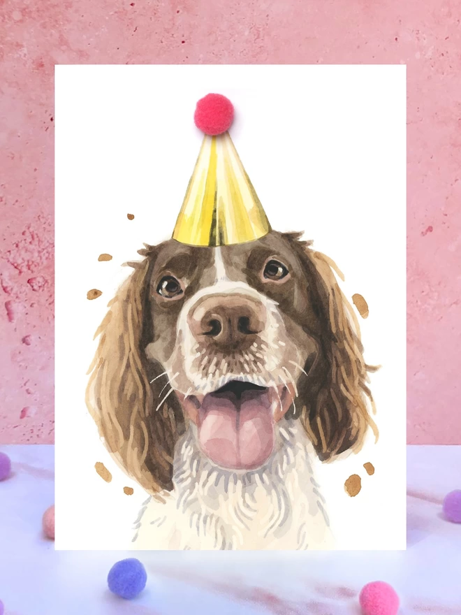 A greeting card featuring a hand painted design of a springer spaniel, stood upright on a marble surface surrounded by pompoms. 