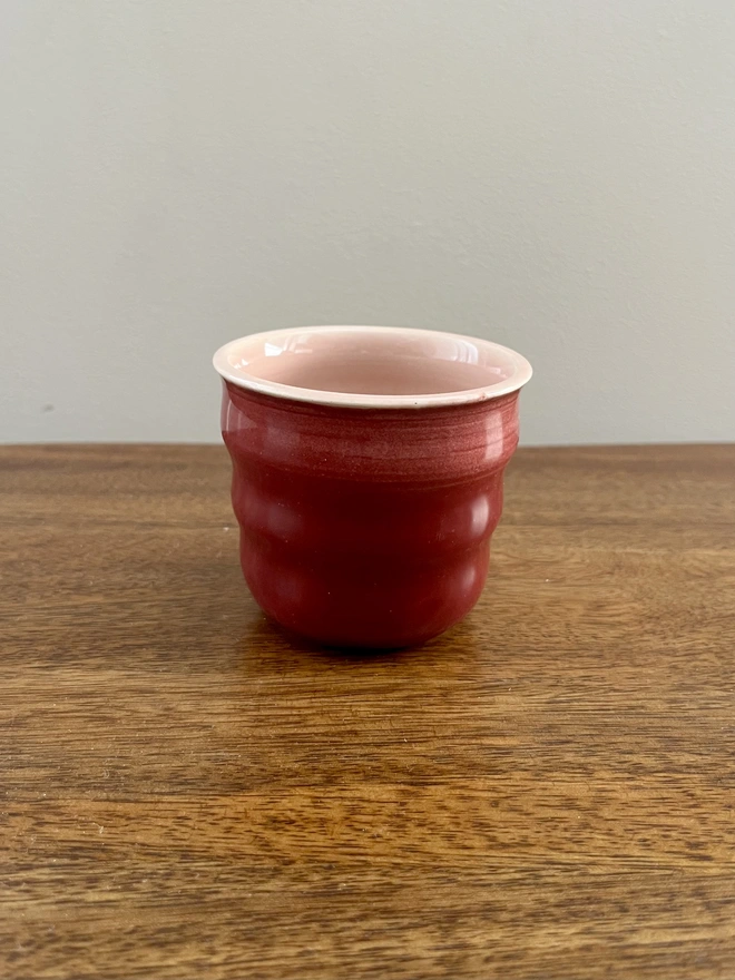 mini ceramic tumbler on a wooden table. the tumbler has a burgundy glaze on the outside and blush pink on the inside and rim. 
