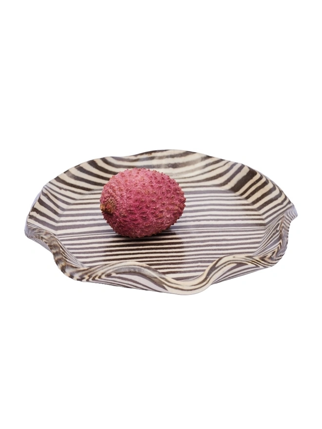 Striped Trinket Dish, a colourful trinket dish sitting on a white surface against a plain backdrop. It is accompanied by some lychees. 