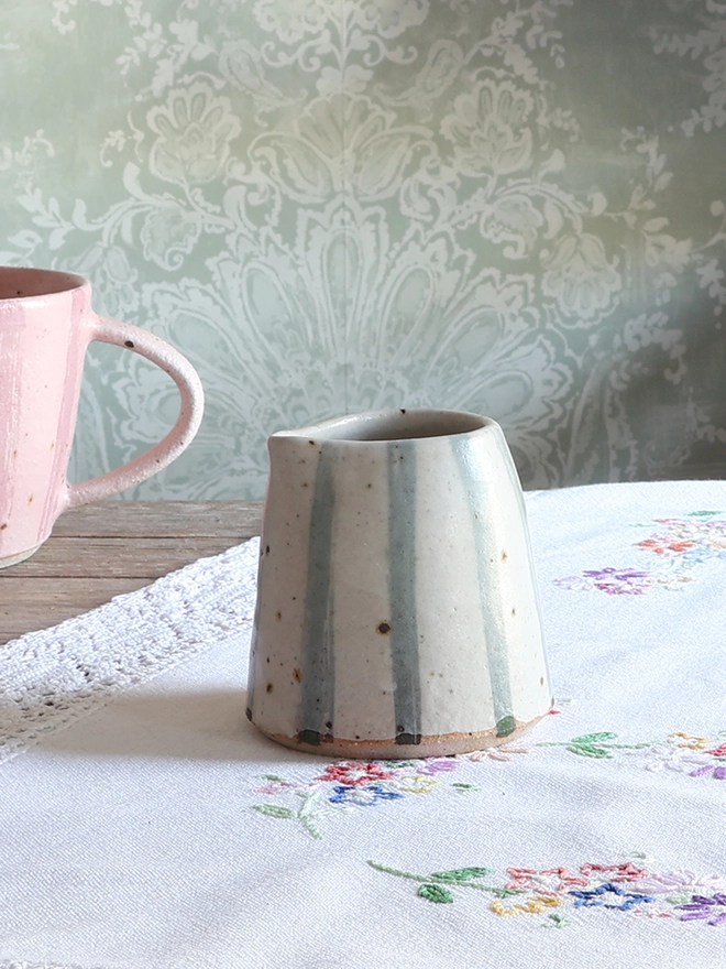 Handmade Ceramic Stoneware Small Jug in Blue and White Stripe sat on a wooden surface covered in a vintage floral tablecloth. A handmade ceramic mug with a pink stripe design sits behind the jug. 