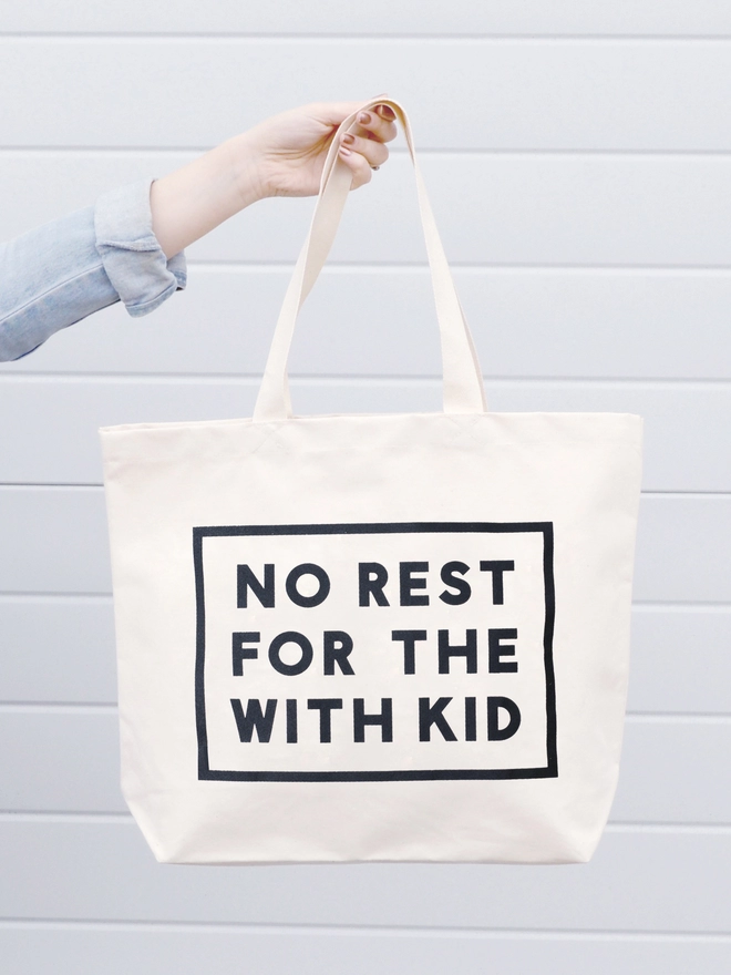 a hand holding out a large natural canvas tote bag bearing the words no rest for the with kid