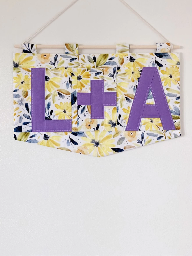 Personalised quilted wall-hanging by Cooper and Fred using a yellow floral pattern and purple lettering.