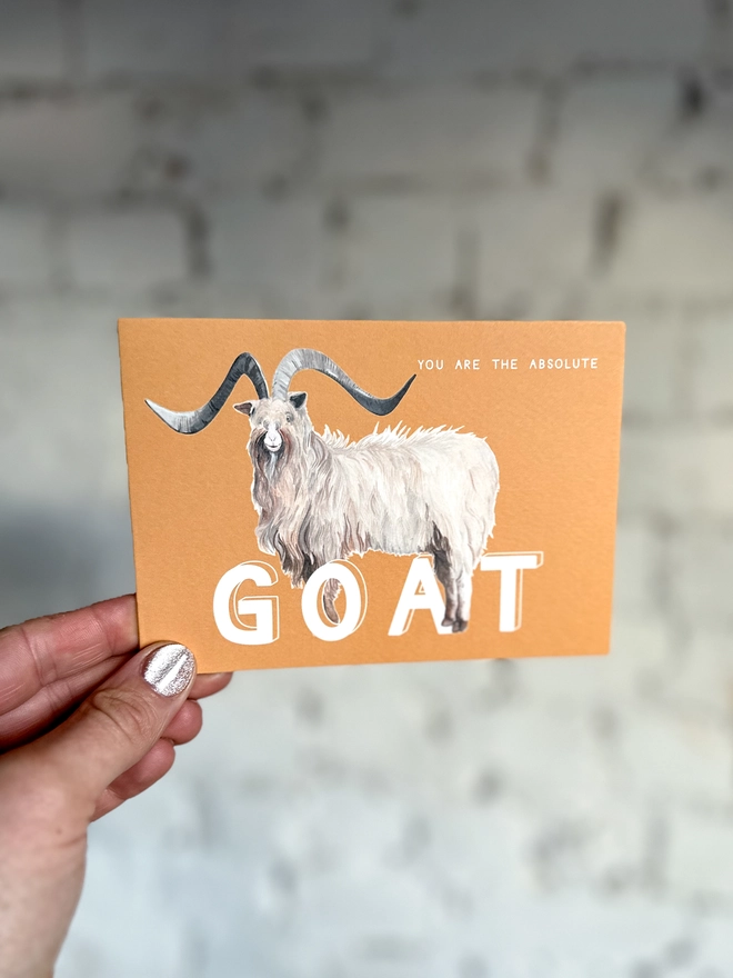 an orangey yellow greetings card featuring an illustration of a bilberry goat with the text “you are the absolute GOAT”