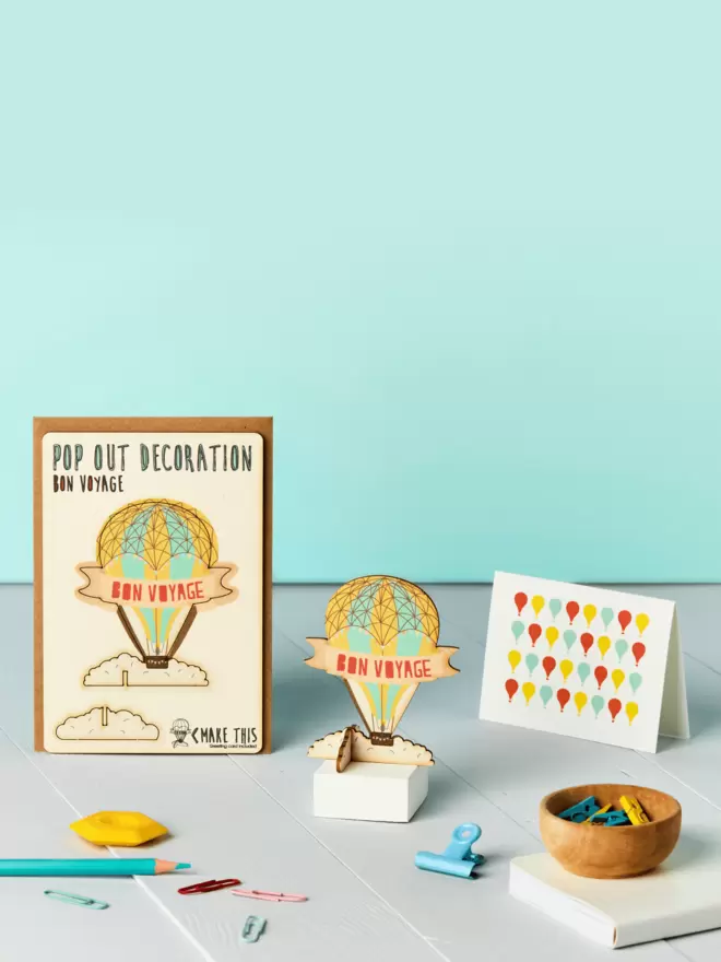 3D bon voyage hot air balloon leaving decoration and hot air balloon pattern card and brown kraft envelope on top of a wooden desk in front of a pale blue coloured background
