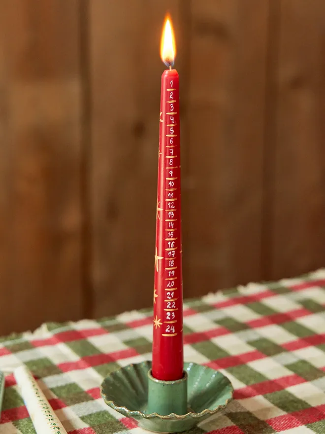 '24 Nights To Christmas' Advent Candle placeed on a checkered red and green tablecloth in a green candlestick holder