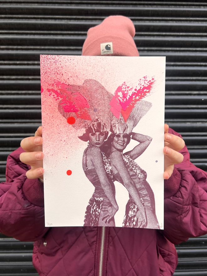 woman artist holding an A4 colourful pink and peach and burgundy print of 2 women in 1920s party clothes with hand painted swooshes and splats, 