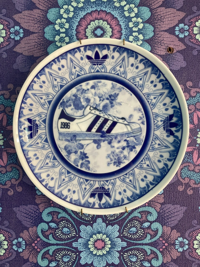 The plate, a blue and white delft style is shown hanging on a wall. It features an Adidas Superstar trainer int he centre.