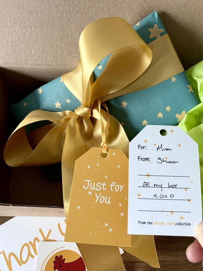 Gift wrapped parcel with blue paper and gold stars and wide golden ribbon in a large bow, in a propped up in a box for posting. Attached yellow gift card says 'Just for You' with hand holding second gift card showing the back with message to recipient. Lime green tissue and business thank you card in background.