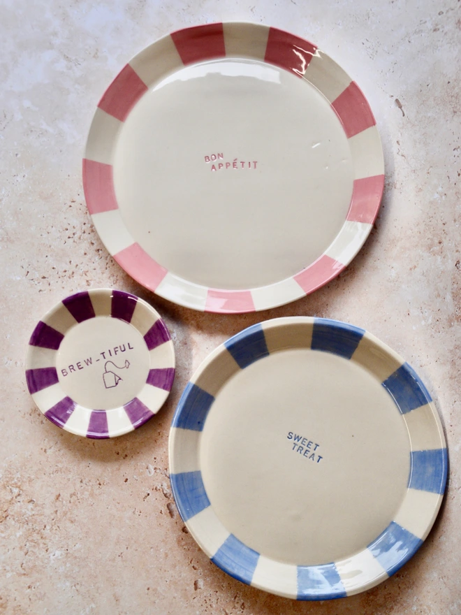 Three handmade ceramic plates with bold striped rims in pink, blue, and purple tones, arranged on a neutral textured surface. Each plate features a playful phrase in the center: “Bon Appétit,” “Sweet Treat,” and “Brew-tiful” with a small teabag illustration.