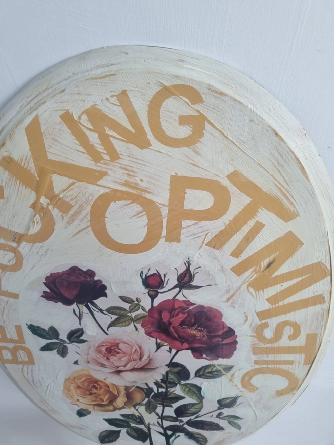 An art panel made from an upcycled table top. The image is vintage roses on a cream background with text that reads 'be fucking optimistic' in mustard yellow..