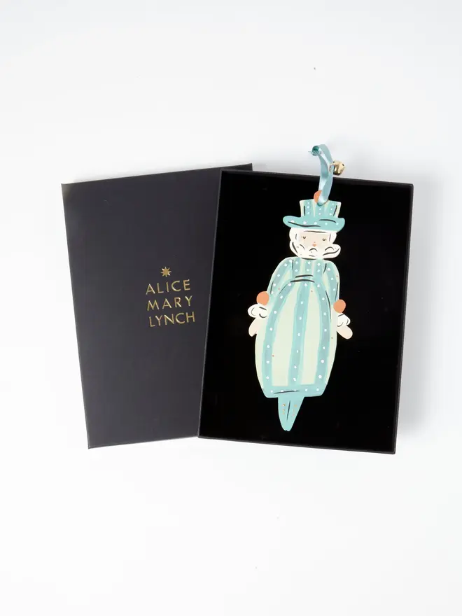 Miss Christmas Tree Dancer. the miss christmas tree dancer hanging ornament sits inside its black presentation box, with the lid open to reveal "alice mary lynch" embossed in gold lettering. the bearded figure in a striped teal and cream costume with polka dots hangs by its teal ribbon and gold bell against the black velvet-lined interior. the branded box gives the piece a polished, gift-ready presentation.