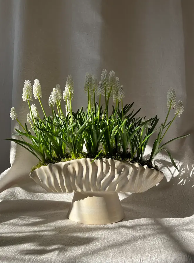 Textured Oval Pedestal Planter Opal, an opal planter sitting on a linen surface. It is accompanied by some plants. 