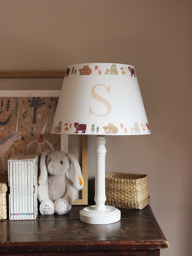 Cozy corner of a girls bedroom, featuring a wooden dresser and white table lamp paired with a personalised kids lampshade adorned with a bears pattern and the letter 'S'. A framed artwork hangs behind the lampshade featuring a unicorn and mythical setting. 