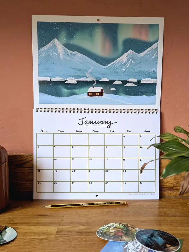 Illustrated double page wall calendar, featuring a cabin against blues and pinks of the northern lights 

