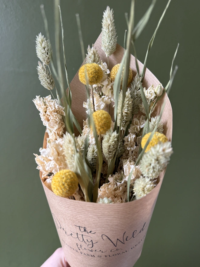 a bunch of dried flowers in bright colours wrapped in black tissue and kraft paper held by a person in a mustard coloured jumper with their left arm against a green wall