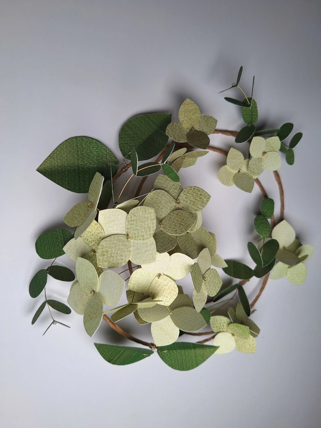 Green hydrangea flower wreath, handmade from paper. There is a cluster of 16 hydrangea blooms on the wreath, which is also adorned with single flowers and two types of dark green leafy stems.