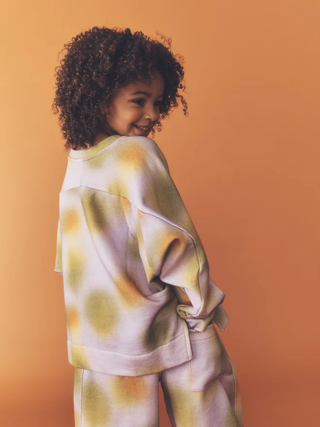 Child looks over their shoulder against a burnt orange backdrop, showing the back and side split of the Studio Koter Riverson top in Hazy Lilac with matching joggers. 