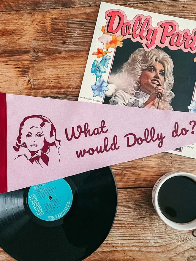 A pastel pink felt pennant with burgundy trim and ties, printed in burgundy the beautiful face of country adn western singer Dolly Parton and the phrase what would Dolly do
