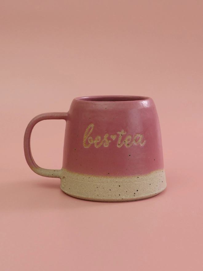 a hot pink handmade pottery mug with bestea saying on the front, sits infront of a pink background