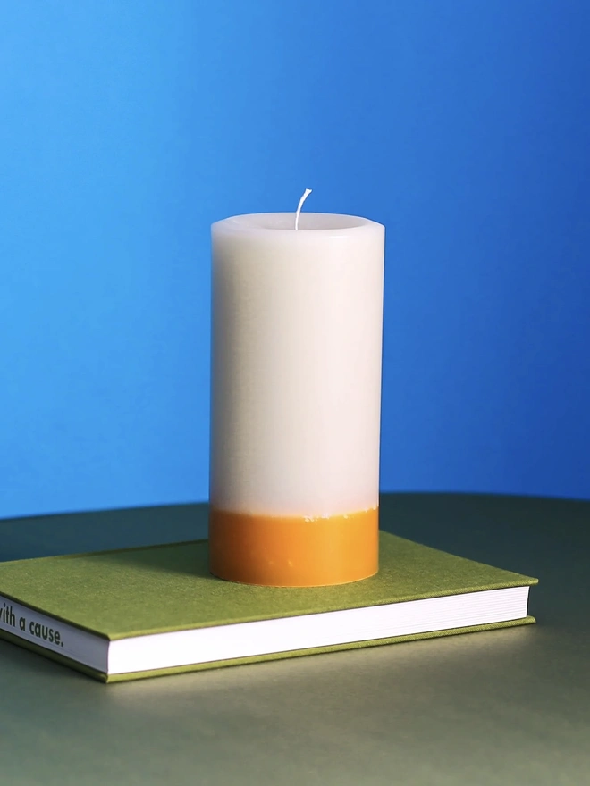 Jasmine, Green Tea & Mulberry Candle (Set Of 1 Or 3 Wicks). a taller jasmine, green tea and mulberry candle sits on a green book against a bright blue background, showing its single wick and cream wax with a golden yellow base. the two-tone colouring and textured, slightly uneven wax surface give the candle a rustic, hand-poured finish. the bold blue background highlights the candle's warm, honeyed tones.