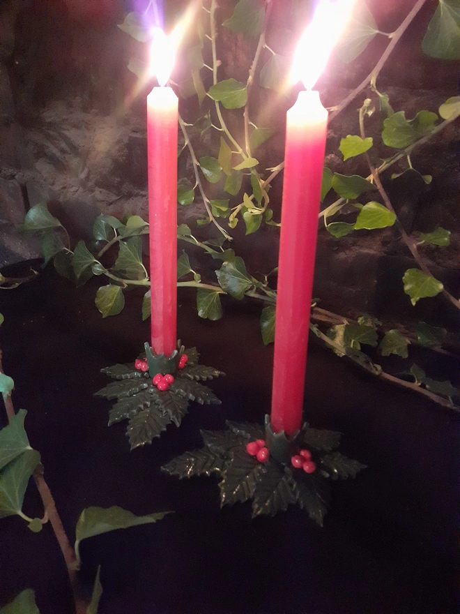 Two green holly leaf candle holders with red berries and lit red taper candles, with trailing ivy in the background.