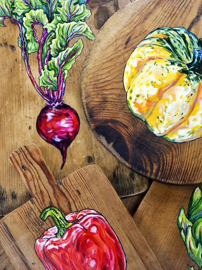 A collection of wooden chopping boards with different handpainted designs including a squash, a red pepper, an artichoke and a beetroot