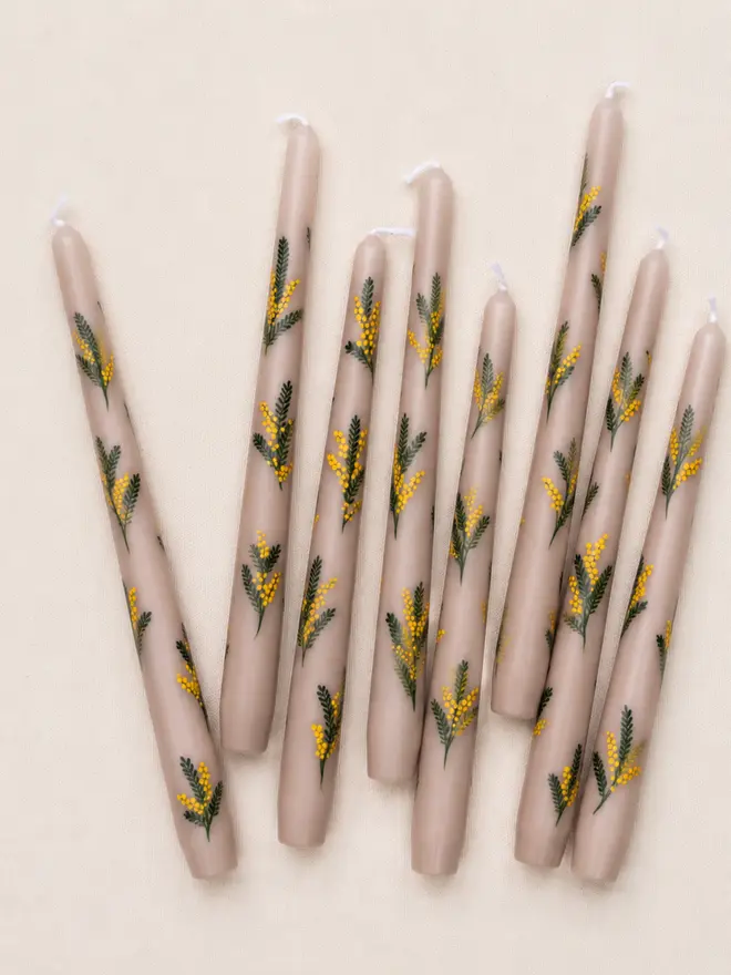 Hand-painted oat-coloured taper candles featuring yellow mimosa flowers and green botanical details, arranged in a relaxed flat lay on natural fabric.