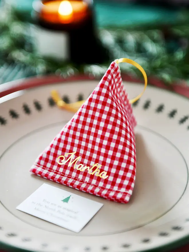 A red gingham fabric fortune cookie with gold personalised name “Martha” sits on a Christmas dinner plate, ready as a handmade festive place setting with a hidden Christmas wish card beneath.