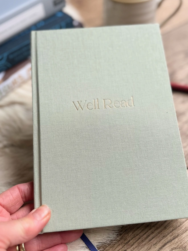 A woman's hand is holding up a pale green linen reading journal. It has been printed with the word Well Read on the front in gold. In the background is a pile of books.