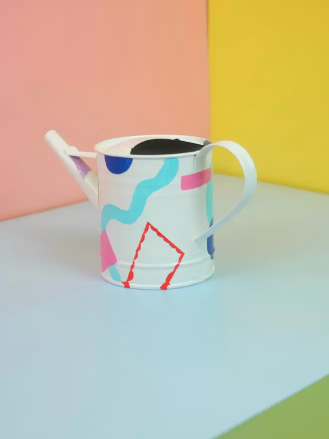 Hand painted watering can by Julie-Anne Pugh. Base colour is white with bring coloured shapes weaving across the body of the can on a yellow and orange background. Colours on shapes include red, violet, peach, light blue, royal blue and green.