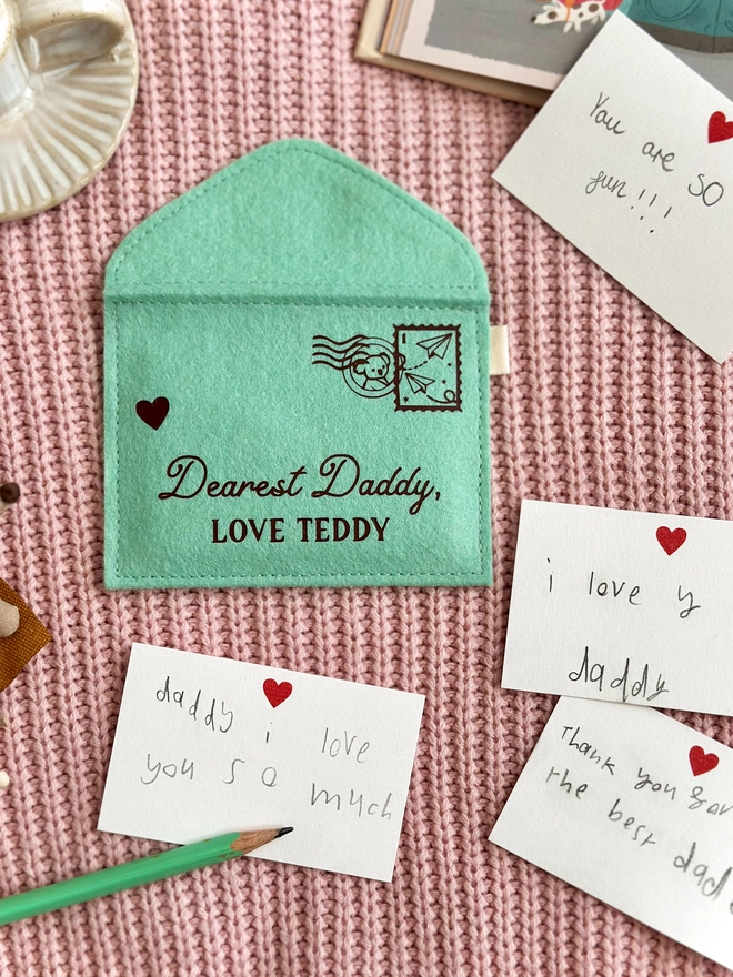 A handmade green felt envelope with the personalisation 'Dearest Daddy, love Maude' and a paper aeroplane stamp with postmark, lays on pink fabric. Notes to Daddy are written on the included heart notecards.