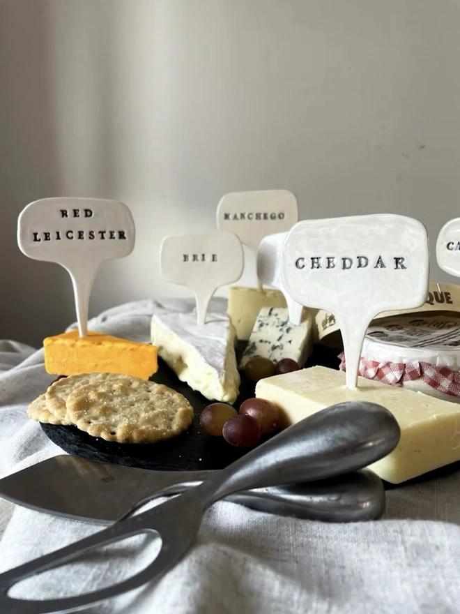 Ceramic Cheese Markers, a group of ceramic cheese markers embedded in a cheese platter. They read: 'Red Leicester, Brie, Manchego, Cheddar'.