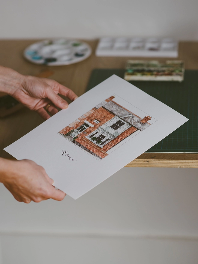 Hands holding a white page with a beautiful edwardian style house in the centre painted in intricate watercolour details. Below black calligraphy lettering reads home
