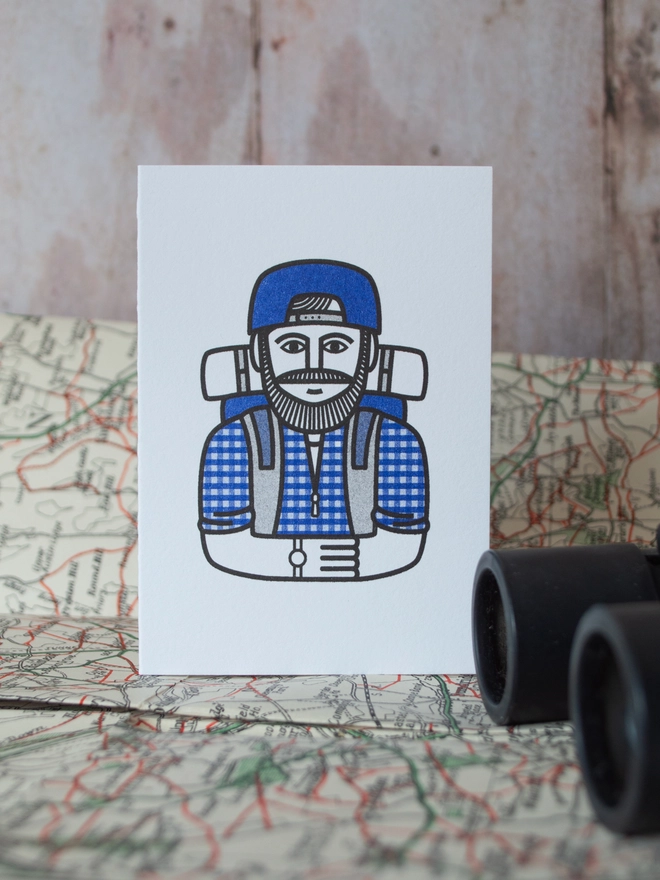 A bold modern illustration of a classic hiker in a blue gingham shirt with cap, rucksack and watch is printed onto a mini greeting card and placed on a vintage map with a pair of binoculars just visible.
