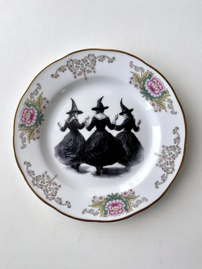 a coven of 3 witches dance hand in hand, the image is victorian in black and white and is printed directly onto an ornate vintage plate