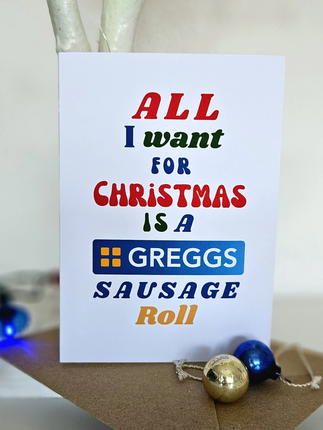 A colourful card Christmas Card that says All I want for Christmas is a Greggs Sausage Roll in retro style fonts on a white background.