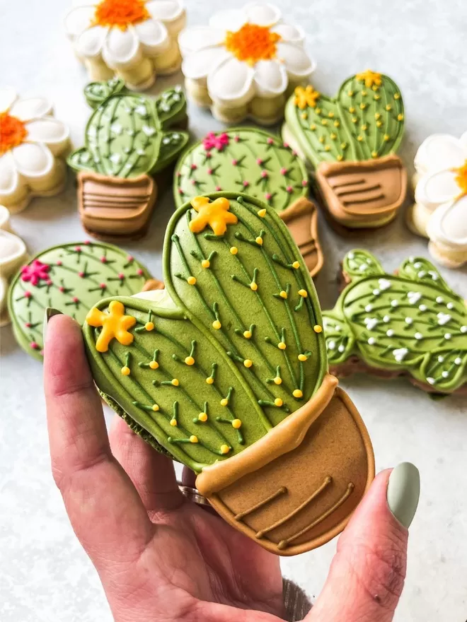 a hand holding a large cactus shaped macaron decorated with yellow flowers with more macaron cactus and daisies in the background