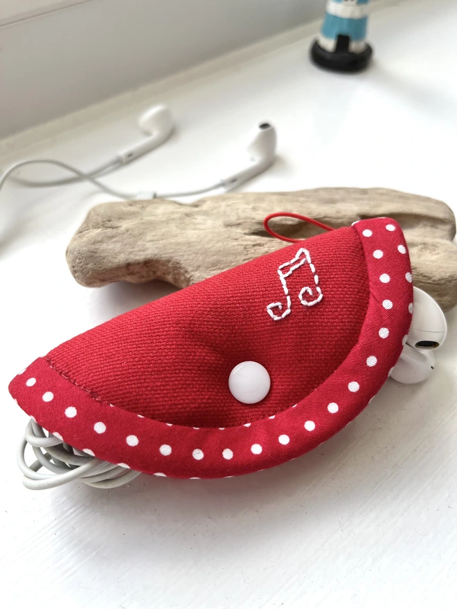 Soft red cotton fold-over earphone case embroidered with musical notes, resting on driftwood.