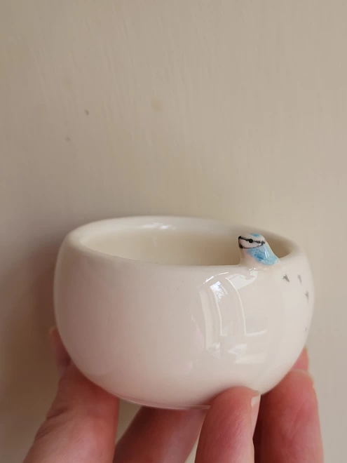 Tiny ceramic bluetit bird is attached to a white pottery tea light holder or trinket dish with tiny bird prints on the side of the pot It is held in a hand